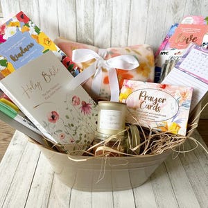May include: A beige basket filled with a variety of gifts, including a Holy Bible, a floral patterned journal, a candle, a blanket, and prayer cards. The basket is filled with shredded paper and has a white ribbon tied around it.