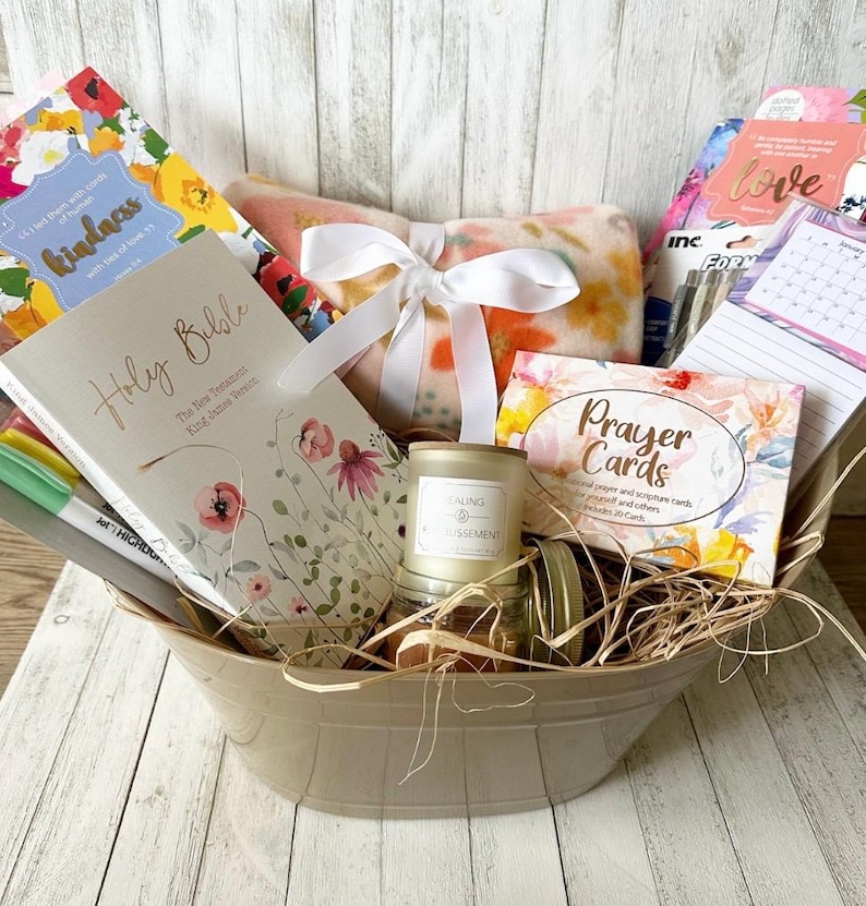 May include: A beige basket filled with a variety of gifts, including a Holy Bible, a floral patterned journal, a candle, a blanket, and prayer cards. The basket is filled with shredded paper and has a white ribbon tied around it.