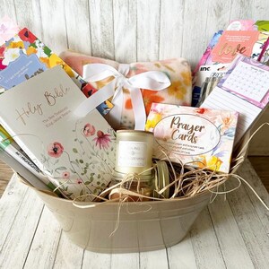 May include: A beige basket filled with a variety of gifts, including a Holy Bible, a prayer card deck, a candle, a blanket, and stationery. The basket is filled with straw and has a white ribbon tied around it.