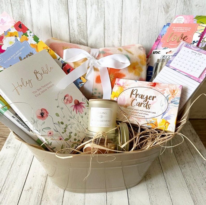 May include: A beige basket filled with a variety of gifts, including a Holy Bible, a prayer card deck, a candle, a blanket, and stationery. The basket is filled with straw and has a white ribbon tied around it.
