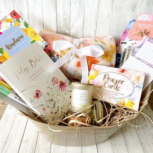 May include: A gift basket filled with items for relaxation and spiritual reflection. It includes a Holy Bible, a prayer card deck, a candle, a throw blanket, and a journal. The basket is lined with straw and has a floral theme.
