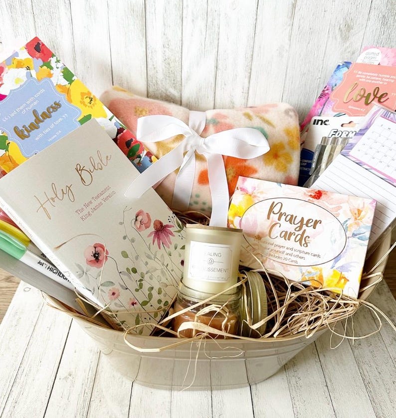 May include: A gift basket filled with items for relaxation and spiritual reflection. It includes a Holy Bible, a prayer card deck, a candle, a throw blanket, and a journal. The basket is lined with straw and has a floral theme.