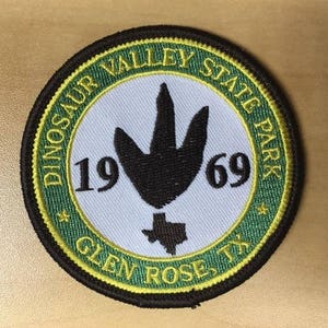 May include: Green and yellow embroidered patch with the text "Dinosaur Valley State Park Glen Rose, TX" and the years "1969" surrounding a black dinosaur footprint.