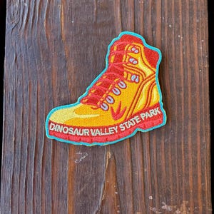May include: An embroidered patch shaped like a hiking boot in shades of yellow and orange with red laces and a red sole. The patch features a bright blue border and the text DINOSAUR VALLEY STATE PARK in white letters along the bottom edge.