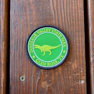 May include: A circular embroidered patch featuring a bright green center with a yellow dinosaur silhouette. The border is black with the text DINOSAUR VALLEY STATE PARK and GLEN ROSE, TX written in yellow, separated by two small yellow stars.
