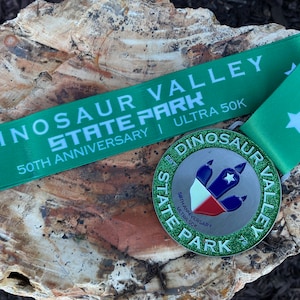 May include: A green ribbon with white text that reads "Dinosaur Valley State Park 50th Anniversary | Ultra 50K". A silver medal with a green glitter finish and the text "Dinosaur Valley State Park" is on top of a brown and green rock.