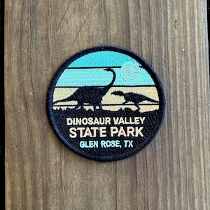 May include: A circular patch featuring a landscape with dinosaurs and the text "DINOSAUR VALLEY STATE PARK GLEN ROSE, TX." The design includes a blue sky, a yellow and black landscape, and a white sun. The patch is black with a stitched border.