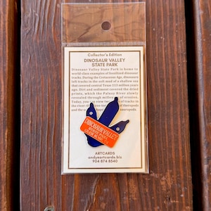 May include: A blue enamel pin shaped like a three-toed dinosaur footprint with an orange banner across the front that reads DINOSAUR VALLEY STATE PARK GLEN ROSE, TX. The pin is mounted on a white card inside a clear plastic sleeve.