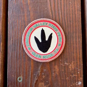 May include: A circular embroidered patch featuring a black dinosaur footprint centered on a cream background. The border is red with a tan outer edge. Green text reads DINOSAUR VALLEY STATE PARK and GLEN ROSE TX, separated by two small green stars.