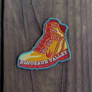 May include: Embroidered patch featuring a hiking boot design in yellow, red, and orange. The boot has red laces and the words "DINOSAUR VALLEY" in white lettering on a red border, outlined in blue.