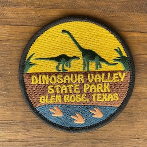May include: Round patch featuring a yellow sky and a brown landscape with two green dinosaurs. The text "DINOSAUR VALLEY STATE PARK GLEN ROSE, TEXAS" is embroidered in gold. A blue water area with dinosaur footprints is at the bottom.