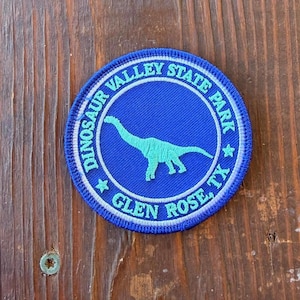 May include: A circular embroidered patch featuring a light blue dinosaur silhouette centered on a dark blue background. The border contains the text DINOSAUR VALLEY STATE PARK and GLEN ROSE, TX in light blue lettering with two small stars.