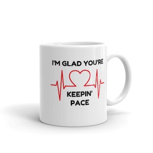 Puede incluir: Taza de cerámica blanca con un corazón rojo y un gráfico de línea de latido del corazón rojo. El texto "I'm glad you're keepin' pace" está impreso en la taza.