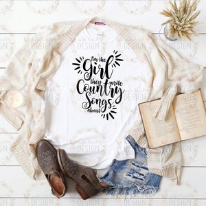 Puede incluir: Camiseta blanca con texto negro que dice "I'm the girl they write country songs about". La camiseta est&aacute; colocada sobre una superficie de madera con una chaqueta blanca de punto, un par de botas marrones y un libro.