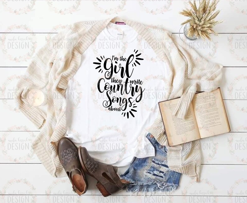 Puede incluir: Camiseta blanca con texto negro que dice "I'm the girl they write country songs about". La camiseta est&aacute; colocada sobre una superficie de madera con una chaqueta blanca de punto, un par de botas marrones y un libro.