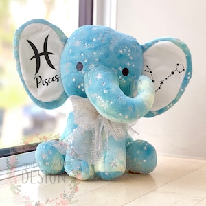 Könnte beinhalten: Ein blauer Plüschelefant mit einem weißen Sternenmuster und einer silbernen Schleife. Die Ohren des Elefanten sind weiß mit schwarzen Tierkreiszeichen, ein Ohr hat das Fische-Symbol und das andere Ohr hat die Fische-Konstellation.