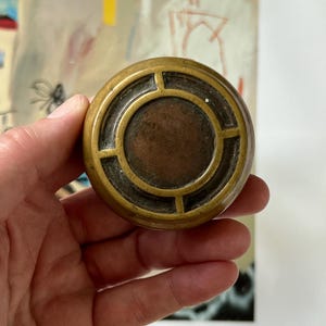 Op de afbeelding: Een ronde, antieke messing deurknop met een cirkelvormig ontwerp. De knop heeft een donkere, verouderde uitstraling met een centraal, verzonken gebied. Het ontwerp heeft een reeks elkaar kruisende lijnen, waardoor een rasterpatroon ontstaat. De knop wordt in een hand gehouden.