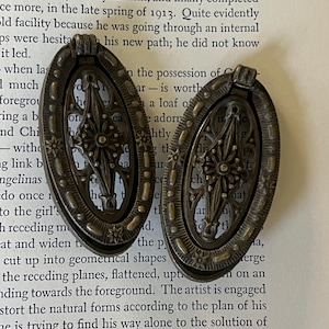 May include: Two oval-shaped metal drawer pulls with a dark bronze finish. Each pull features a hinged drop handle with an intricate, openwork starburst design in the centre, surrounded by a decorative border with small embossed star details.