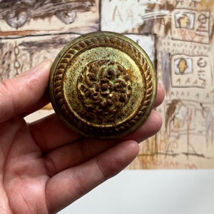 Pode incluir: Puxador de armário antigo em latão com um design circular detalhado. O puxador apresenta um padrão floral central rodeado por uma borda em forma de corda. O acabamento tem uma aparência envelhecida, sugerindo ferragens vintage ou recuperadas.