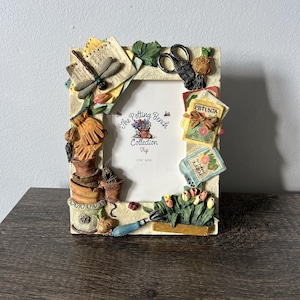 Può includere: Una cornice decorativa a tema giardinaggio. La cornice è beige e presenta una libellula, attrezzi da giardinaggio e bustine di semi di fiori. La cornice ha il testo "The Potting Bench Collection". L'apertura per foto è di 12,7 cm x 17,8 cm.