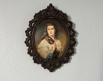 Puede incluir: Retrato ovalado vintage de una mujer con cabello largo, enmarcado en metal decorativo color bronce. El retrato presenta a una mujer con un vestido blanco con un acento azul. El marco tiene un diseño ornamental de volutas.