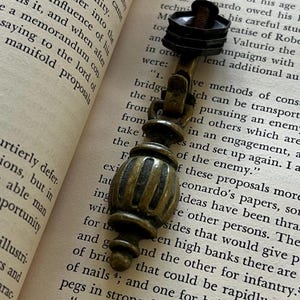 May include: A brass drop pull handle featuring a vertical, ribbed, spindle-shaped body with a rounded finial at the bottom. The hardware includes a hinged bail mechanism and a dark, hexagonal nut attached to a threaded metal bolt at the top end.