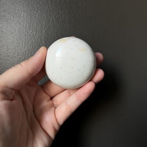 Peut inclure: Une pierre blanche et ronde avec de subtiles marques beiges. La surface lisse et polie reflète la lumière, suggérant un matériau naturel. La pierre est tenue dans une main sur un fond sombre.