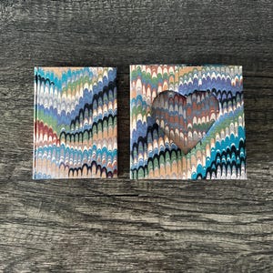 May include: Two small, rectangular notebooks with a marbled paper design in shades of blue, green, and brown. One notebook is whole, while the other has a heart-shaped cutout. The notebooks are on a dark wood surface.