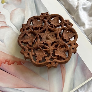 May include: A round, intricately carved wooden decorative piece featuring a floral and leaf design. The brown wood has a detailed, openwork pattern with a central flower and surrounding leaves. The piece is likely a coaster or decorative element.