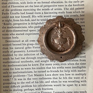 May include: A circular bronze-toned metal drawer pull featuring a scalloped edge and a central embossed urn design. The hardware includes a hinged ring handle and a small mounting hole at the bottom, positioned over a page of printed text.