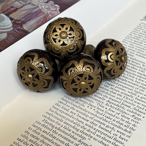May include: Four spherical drawer knobs featuring a dark brown, glossy finish with an intricate, raised gold-toned metal overlay. The metal design consists of a central flower motif surrounded by symmetrical, curved, and leaf-like patterns.