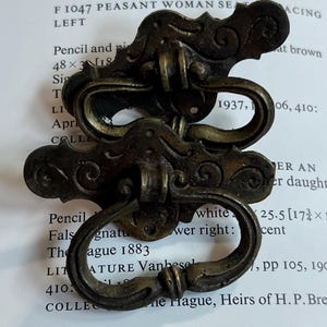 May include: Two antique-style brass drawer pulls featuring ornate, scrolled backplates and swinging drop handles. The metal has a dark, aged patina with visible wear, showcasing intricate decorative detailing on the mounting plates.