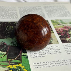 May include: A polished, spherical object with a rich, dark brown color and visible wood grain patterns. The orb appears smooth and reflective, suggesting a decorative or collectible item. The background includes a printed page with text and images.