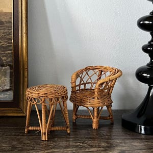 May include: A miniature wicker chair and stool set. The chair has a rounded back and armrests, while the stool is a small, round, four-legged design. Both pieces are made of light brown woven wicker.