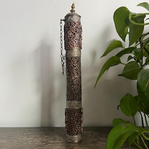 May include: A tall, ornate bronze-colored metal container with intricate cut-out designs. The container has a decorative top and bottom with a chain attached to the lid. The container is cylindrical and stands upright.
