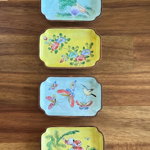 Puede incluir: Cuatro platos rectangulares de cerámica con diseños pintados a mano. Los platos presentan imágenes de verduras, flores, mariposas, un pájaro y una persona. Cada plato tiene un fondo claro y un borde marrón. Los platos se exhiben sobre una superficie de madera.