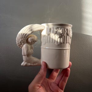 Könnte beinhalten: Cremefarbene Keramiktasse mit einem Griff in Form eines Engels. Der Engel kniet mit gefalteten Händen im Gebet, und die Tasse hat ein strukturiertes Design. Die Tasse wird in einer Hand vor einem neutralen Hintergrund gehalten.