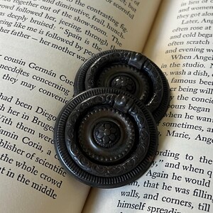 May include: Two circular dark bronze drawer pulls with a swinging ring handle. Each pull features a central floral motif surrounded by a decorative border of repeating heart shapes and a beaded edge, resting on an open book page.