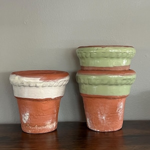 May include: Three terracotta flower pots with a distressed finish. One pot has a white band, while the other two have a green band. The pots are arranged on a dark wooden surface against a gray background.