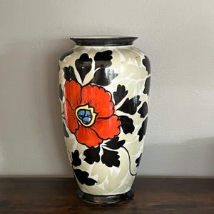 Peut inclure: Un vase décoratif en céramique avec un motif de fleur de pavot rouge vif. Le vase présente un fond crème avec des motifs de feuilles noires et vert clair. Le pavot a un centre bleu. Le vase a un bord et une base noirs.