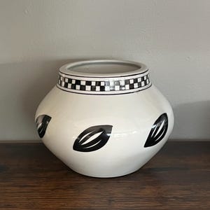 May include: A white ceramic vase with a rounded body and a checkered black and white band around the rim. The vase features three black leaf-shaped designs. The vase is displayed on a wooden surface.