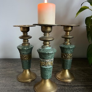 May include: Three candlestick holders featuring a combination of polished brass bases and tops with a central green marble-like stone section. Each holder includes a decorative brass band with embossed floral patterns. A single lit pillar candle sits on the center holder.