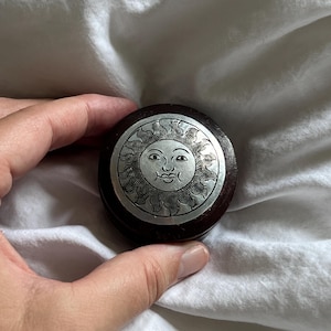 Könnte beinhalten: Eine runde Holzbox mit einem silberfarbenen Deckel, verziert mit einem gravierten Sonnendesign. Die Sonne hat ein lächelndes Gesicht und Strahlen. Die Box befindet sich vor einem weißen, strukturierten Hintergrund.