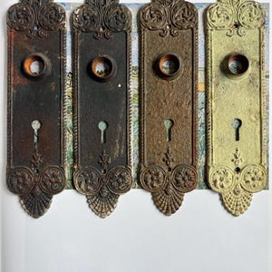 May include: Four antique door plates in varying shades of brown and green. Each plate has a keyhole, a circular opening, and ornate floral and shell designs. The plates are arranged vertically against a colorful background.