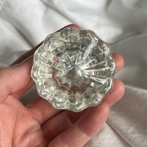 May include: Clear glass door knob with a round, fluted design. The knob is held in a hand against a white background. The glass reflects light, creating a sparkling effect. This vintage-style knob is a decorative hardware element.