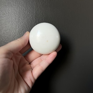 Pode incluir: Um objeto esférico branco e liso. O item redondo tem um acabamento brilhante e parece ser feito de cerâmica ou material semelhante. O objeto é segurado em uma mão contra um fundo escuro.