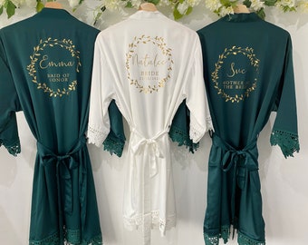 May include: Three satin robes with lace trim, one white and two hunter green. The robes have gold foil lettering with a floral wreath design. The white robe says "Natalie Bride 23.12.2021". The green robes say "Emma Maid of Honor" and "Sue Mother of the Bride".