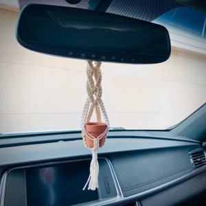 May include: A white macrame plant hanger with a small terracotta pot hanging from the rearview mirror of a car.