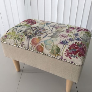 May include: A rectangular footstool with a floral design top in red, purple, and green on a cream base. The sides are light beige fabric, trimmed with silver studs. It has four wooden legs. The dimensions are approximately 40cm x 30cm.