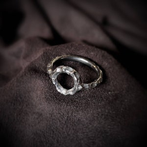 May include: A silver ring with a textured, circular design. The ring is open on one side, creating a unique and modern look.